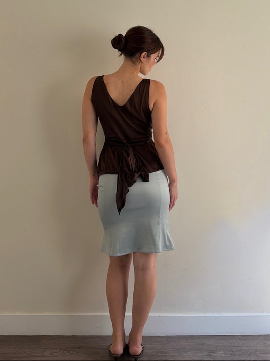 Woman wearing a brown top and light gray skirt against a plain wall.