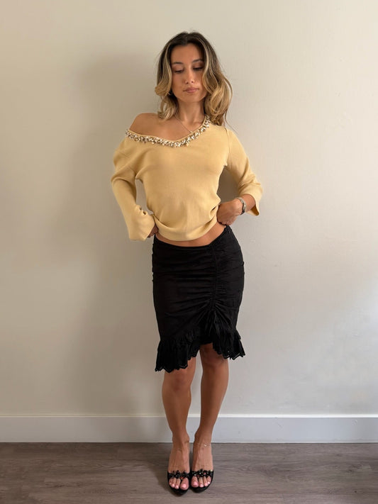 A woman modeling a pale yellow off-the-shoulder knit top with a beaded neckline and a black ruched ruffled midi skirt, standing against a plain beige wall.