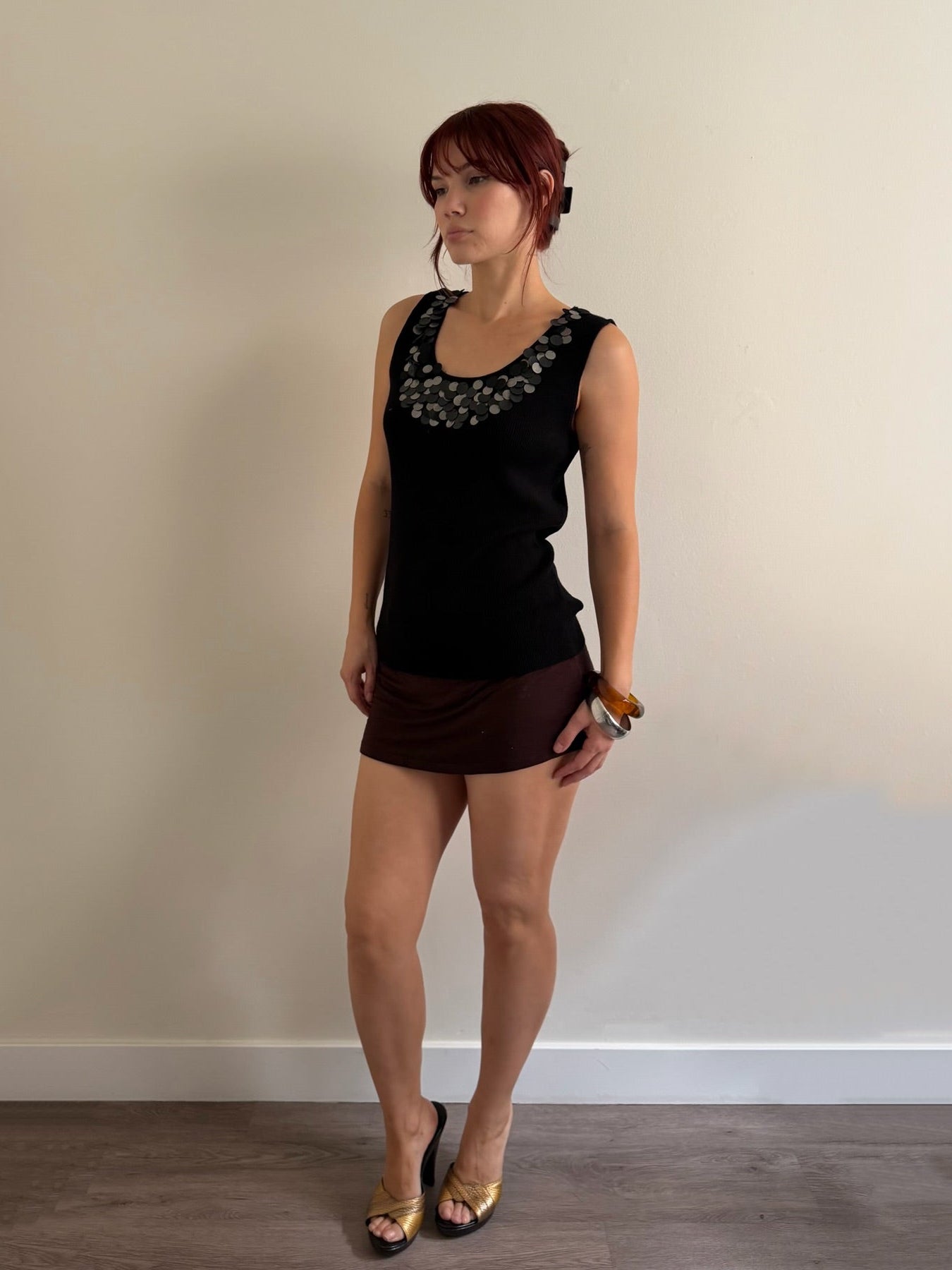 A woman with red hair stands against a plain beige wall wearing a black sleeveless top with a decorative neckline and a dark brown mini skirt, paired with black and gold heels.