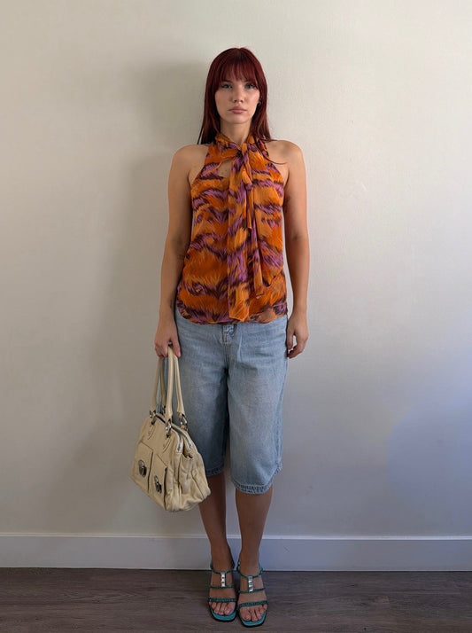 Woman wearing a patterned top and denim shorts against a plain wall.