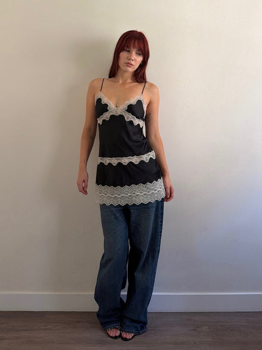 Woman wearing a black and white lace top with blue jeans against a plain wall.