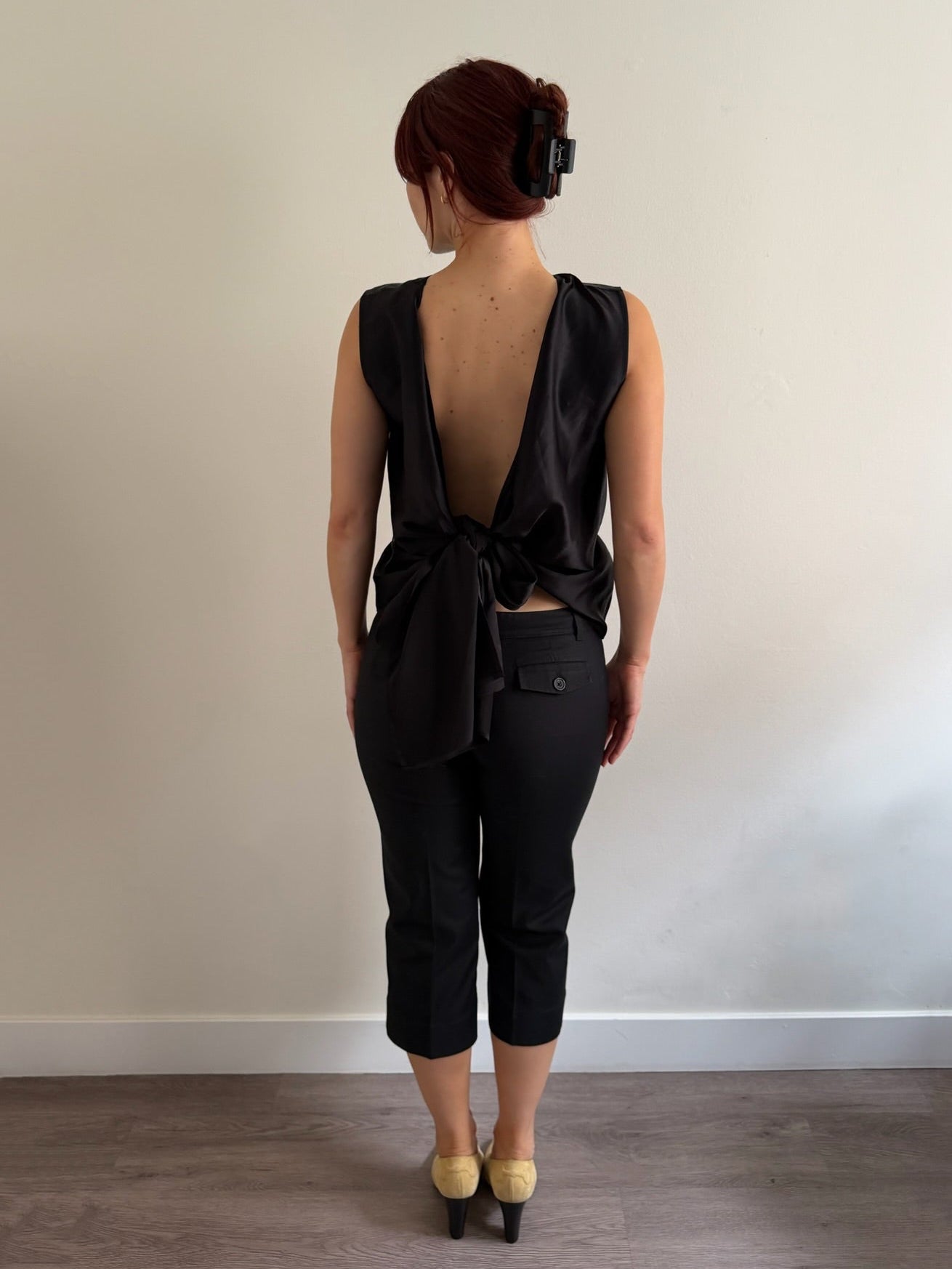 A woman stands with her back to the camera wearing a black satin-like sleeveless top with a deep open back and front knot detail, paired with black cropped pants and beige heels, against a plain off-white wall.