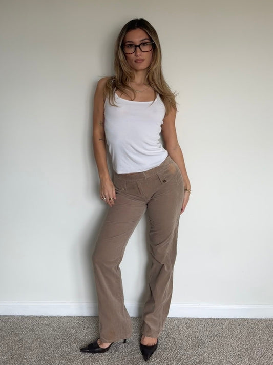 Woman wearing a white tank top and beige pants against a plain wall.