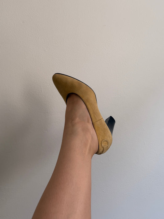 A person's foot wearing a tan suede high-heeled shoe with a black pointed toe, shown against a plain off-white background.
