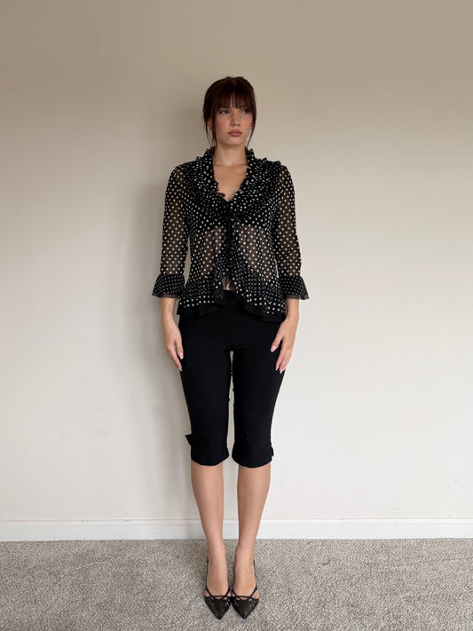 Person wearing a black patterned blouse and black pants against a plain wall.