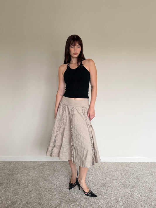 Woman wearing a black top and beige skirt standing against a plain wall.