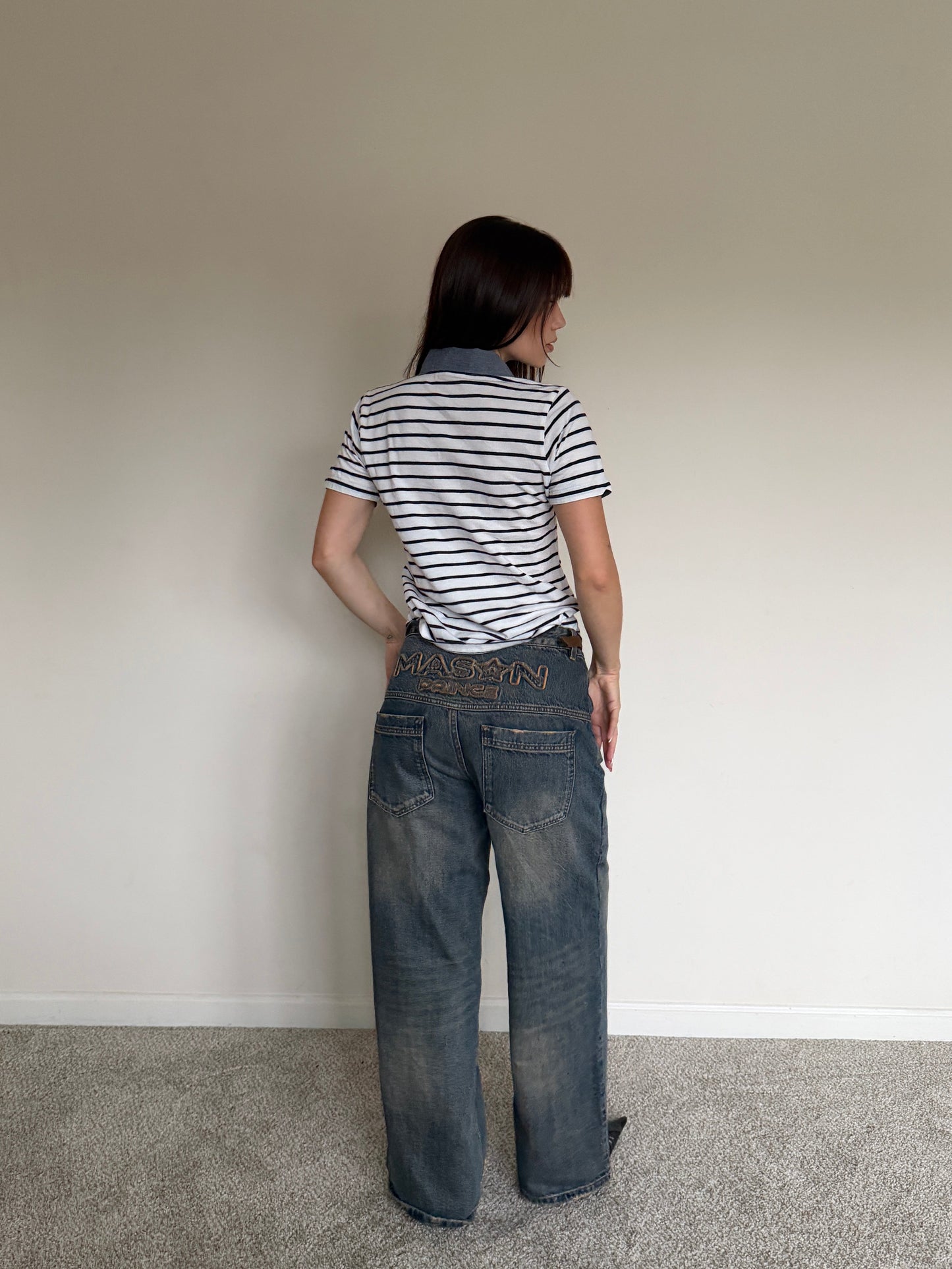 Person wearing a striped shirt and jeans standing against a plain wall.