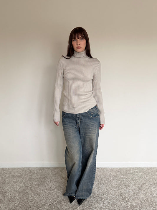 Person wearing a light gray turtleneck sweater and blue jeans against a plain wall.
