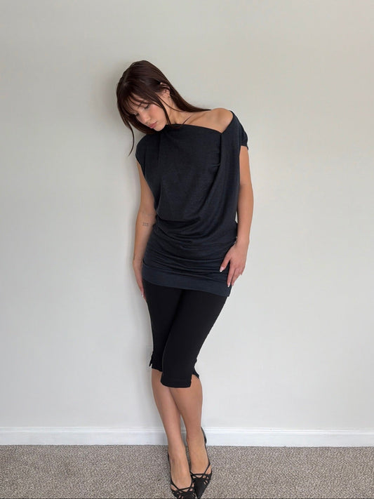 Woman wearing a black off-shoulder top and black pants against a plain white wall.