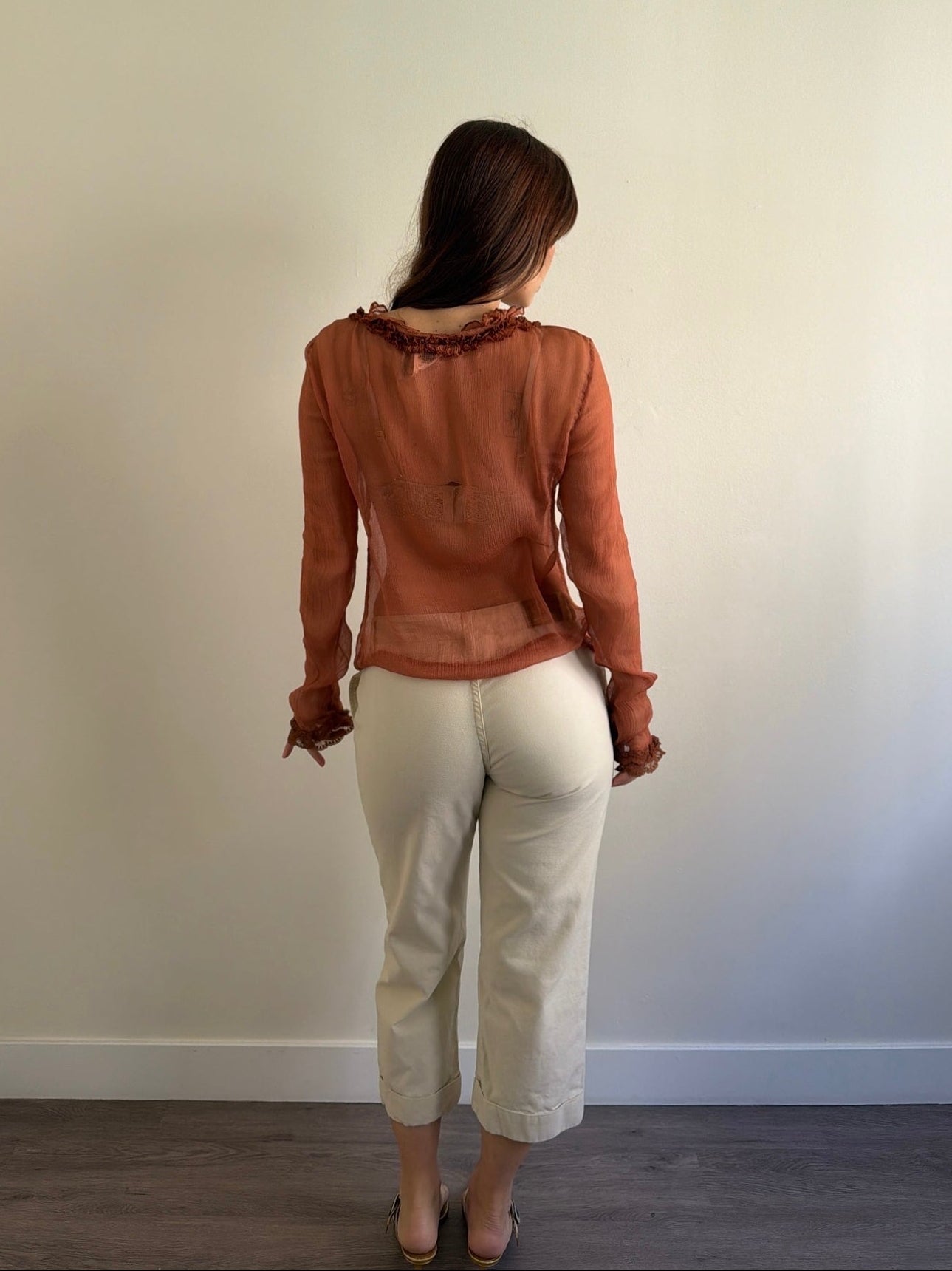 Person wearing a brown blouse and white pants standing against a plain wall.