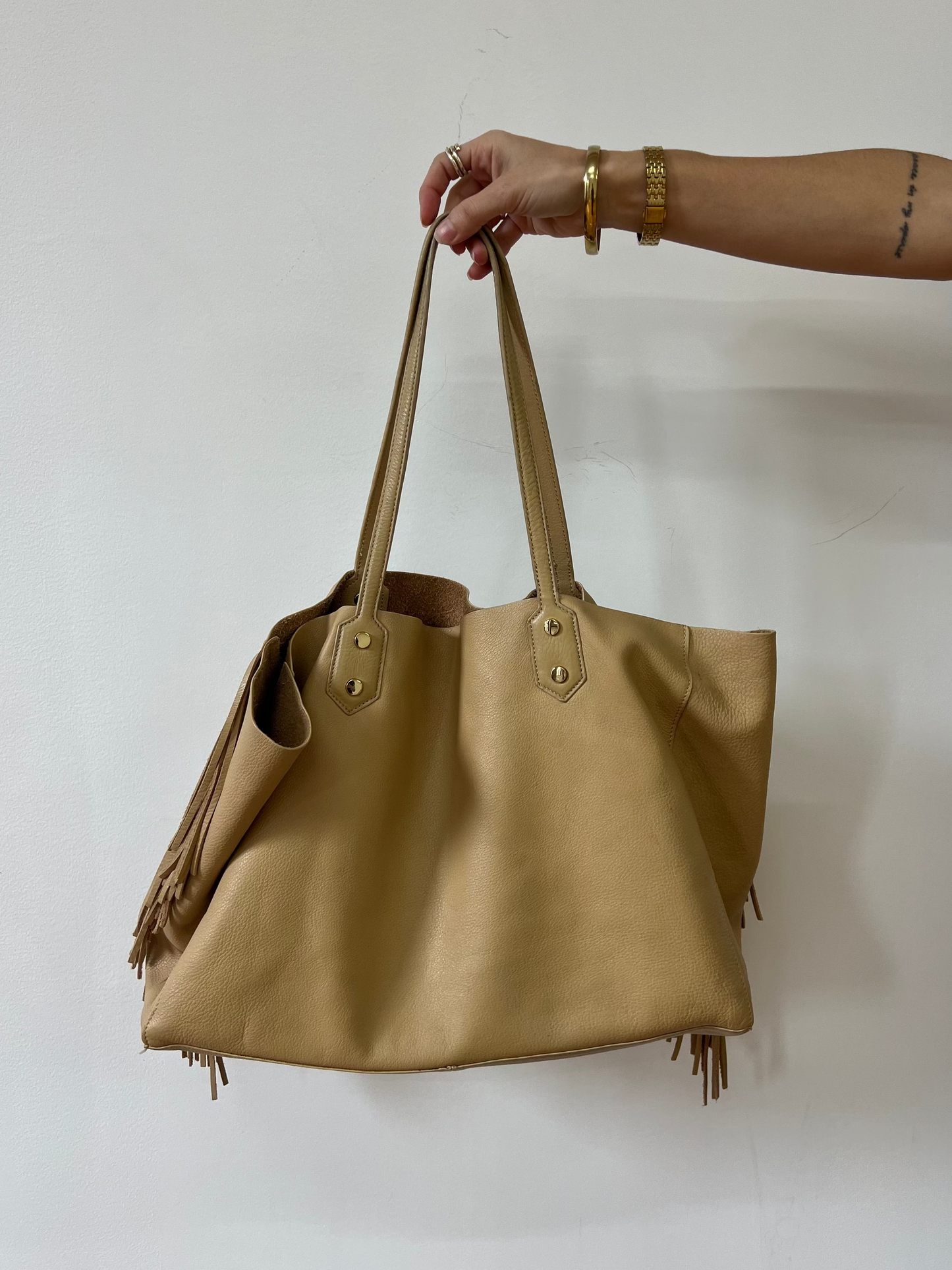 Beige handbag with fringe details held by a hand against a white background