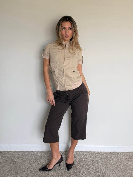 Woman wearing a beige short-sleeve shirt and black pants against a plain wall.