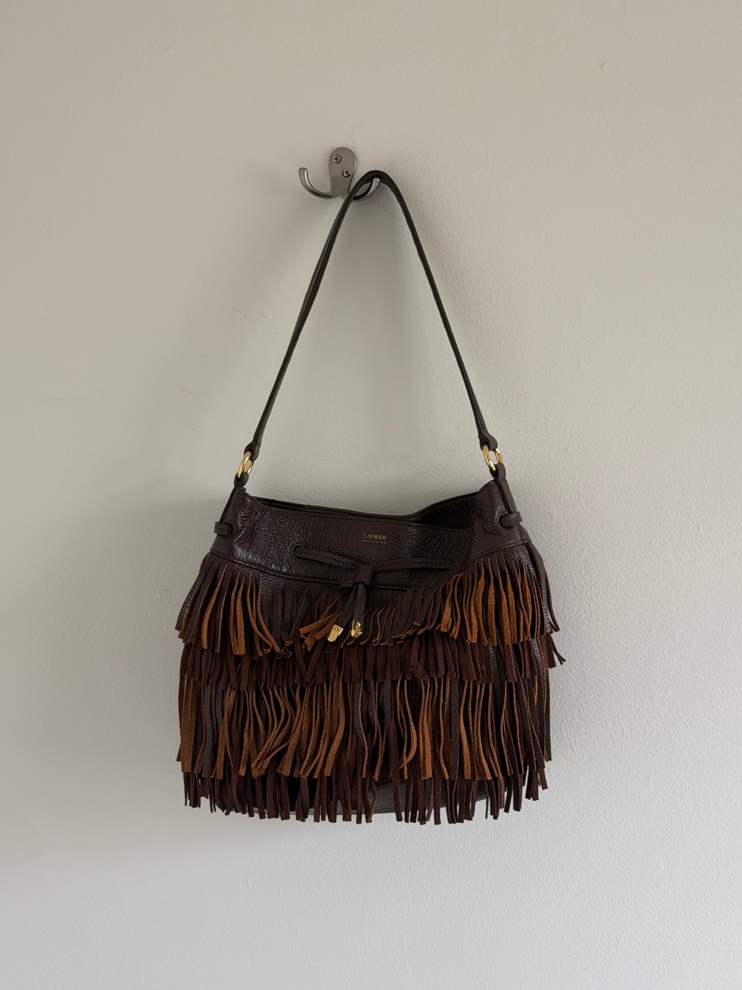 Brown leather handbag with fringe details hanging on a hook against a white wall.