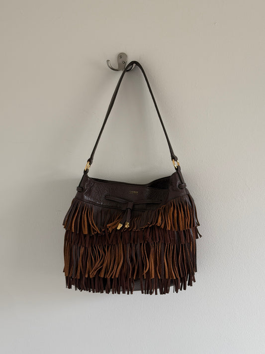 Brown leather handbag with fringe details hanging on a hook against a white wall.