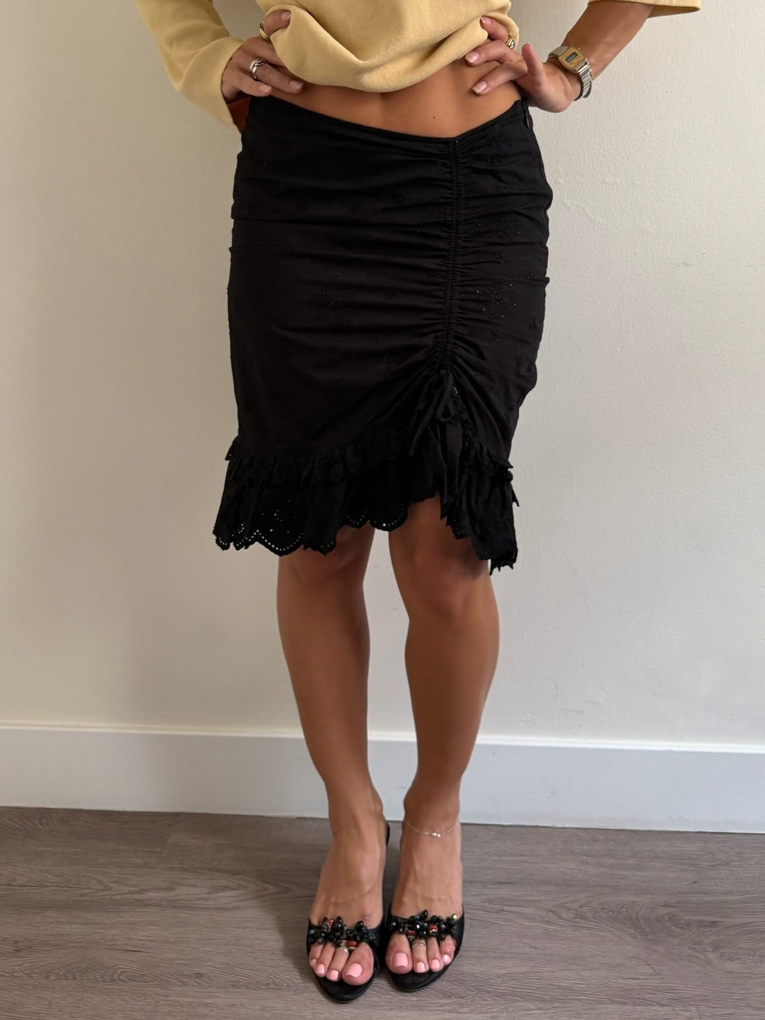 A woman modeling a pale yellow off-the-shoulder knit sweater with a beaded neckline and a black ruched mini skirt, paired with black heeled sandals, standing against a plain beige wall.