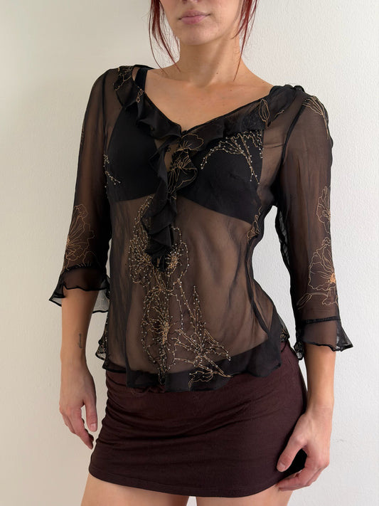 A woman wearing a sheer black ruffled blouse with gold floral embroidery over a black bralette, paired with a dark brown mini skirt, against a plain off-white wall.