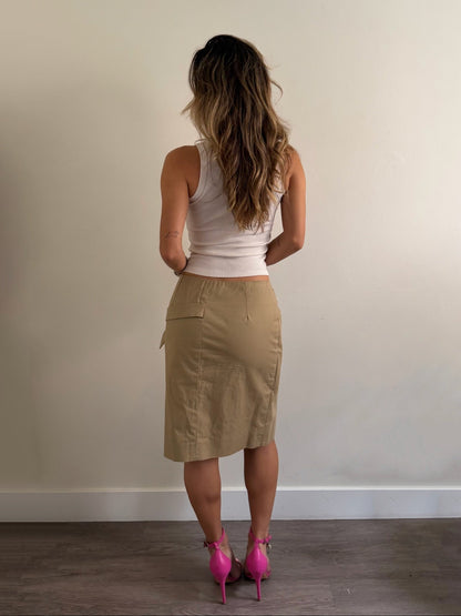 A woman is shown from behind wearing a beige knee-length skirt, a white sleeveless top, and bright pink high heels, standing against a plain off-white wall.