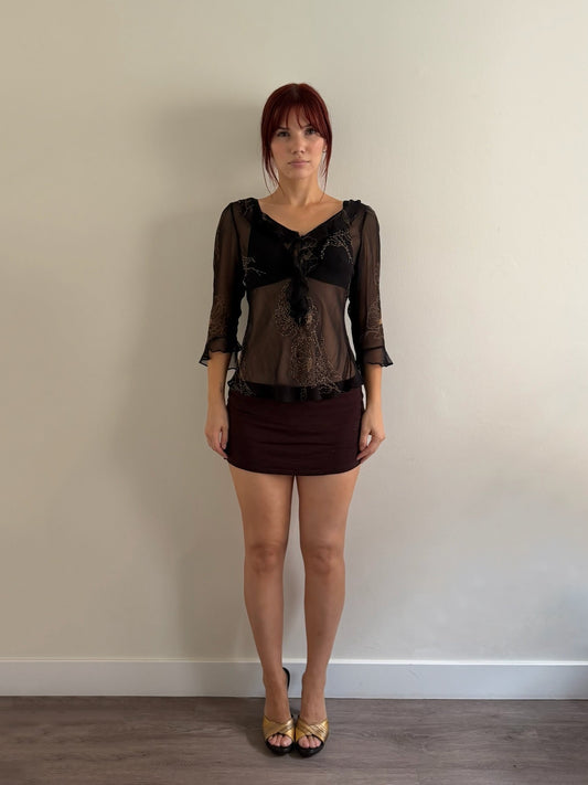 A woman with red hair stands against a plain beige wall wearing a sheer black lace top with three-quarter sleeves and a dark brown mini skirt.