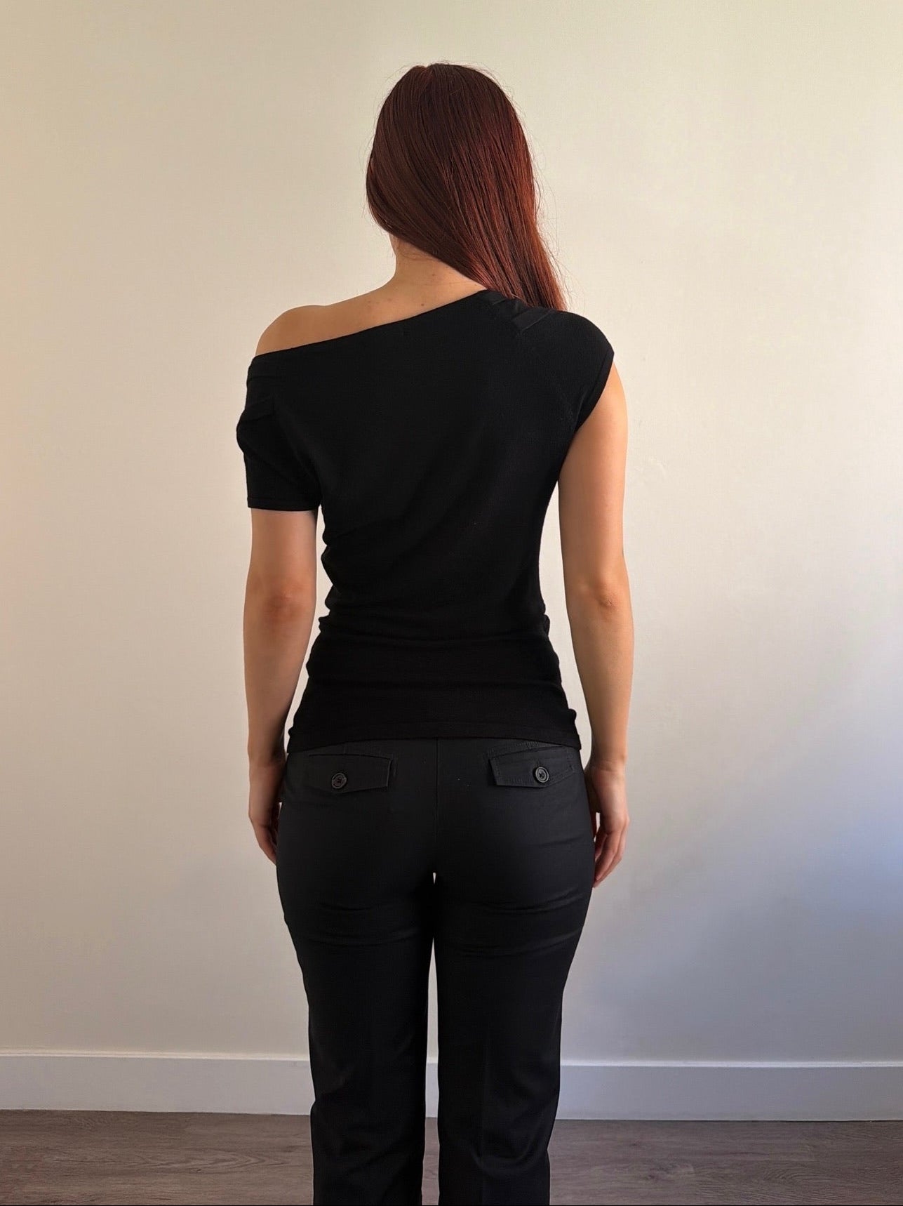 Person wearing a black off-shoulder top and black pants against a plain wall.