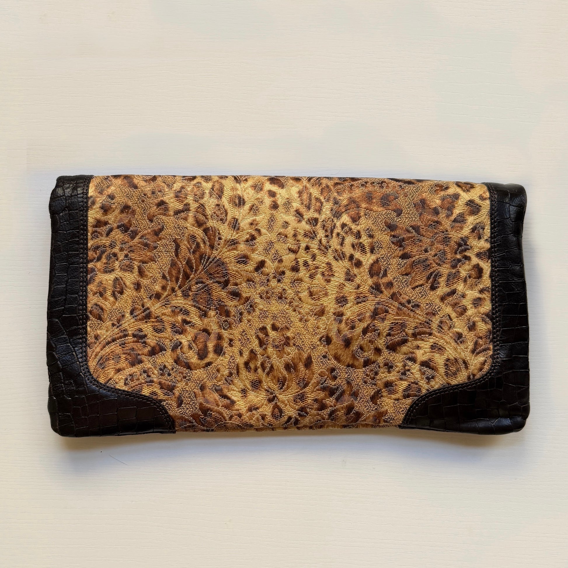 A rectangular clutch wallet featuring a central leopard print panel in beige and brown, framed by black crocodile-embossed leather borders on a light off-white background.