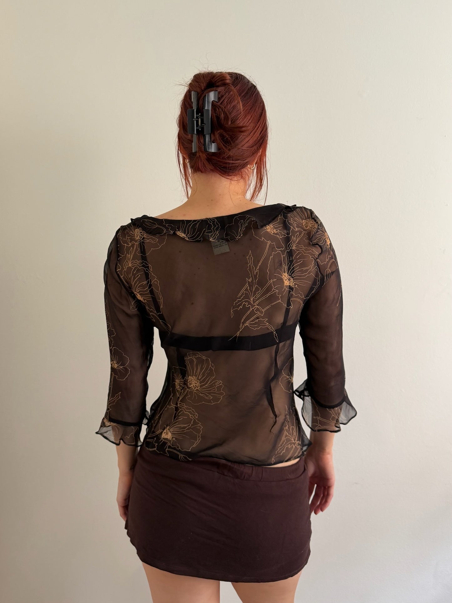 A woman viewed from behind wearing a sheer dark brown floral mesh blouse with black lace trim and a dark brown mini skirt, standing against a plain off-white wall.