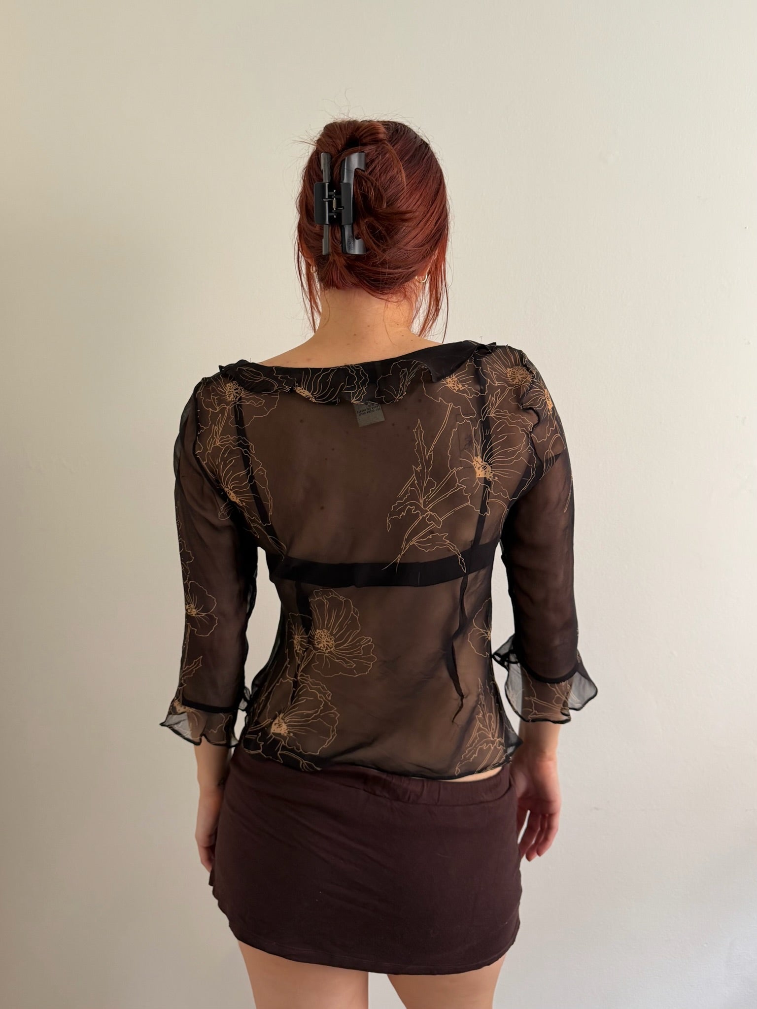 A woman viewed from behind wearing a sheer dark brown floral mesh blouse with black lace trim and a dark brown mini skirt, standing against a plain off-white wall.