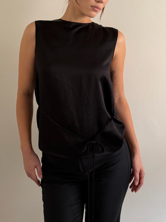 A woman wearing a sleeveless black satin-like top with a draped, gathered waist and tie detail, paired with black pants, against a plain light beige wall.