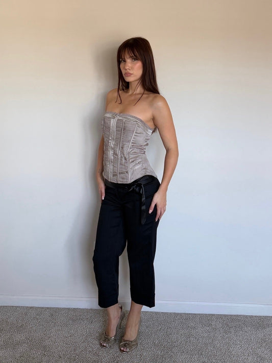 Woman wearing a strapless metallic corset top and black pants against a plain wall.