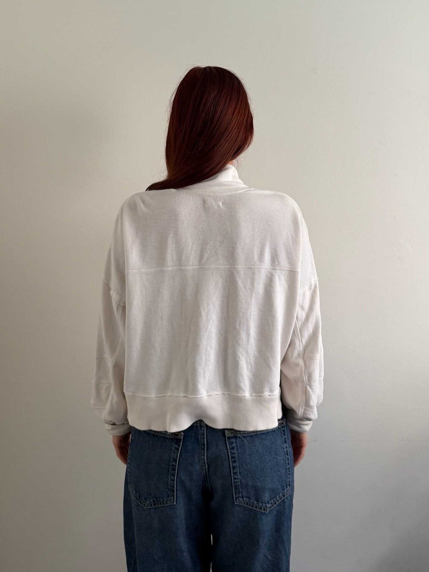 A person viewed from behind wearing a white cropped sweatshirt and blue jeans against a plain light gray wall.