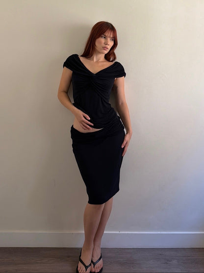 A woman with red hair wearing a black off-the-shoulder ruched midi dress and black flip-flop sandals, standing against a plain beige wall.