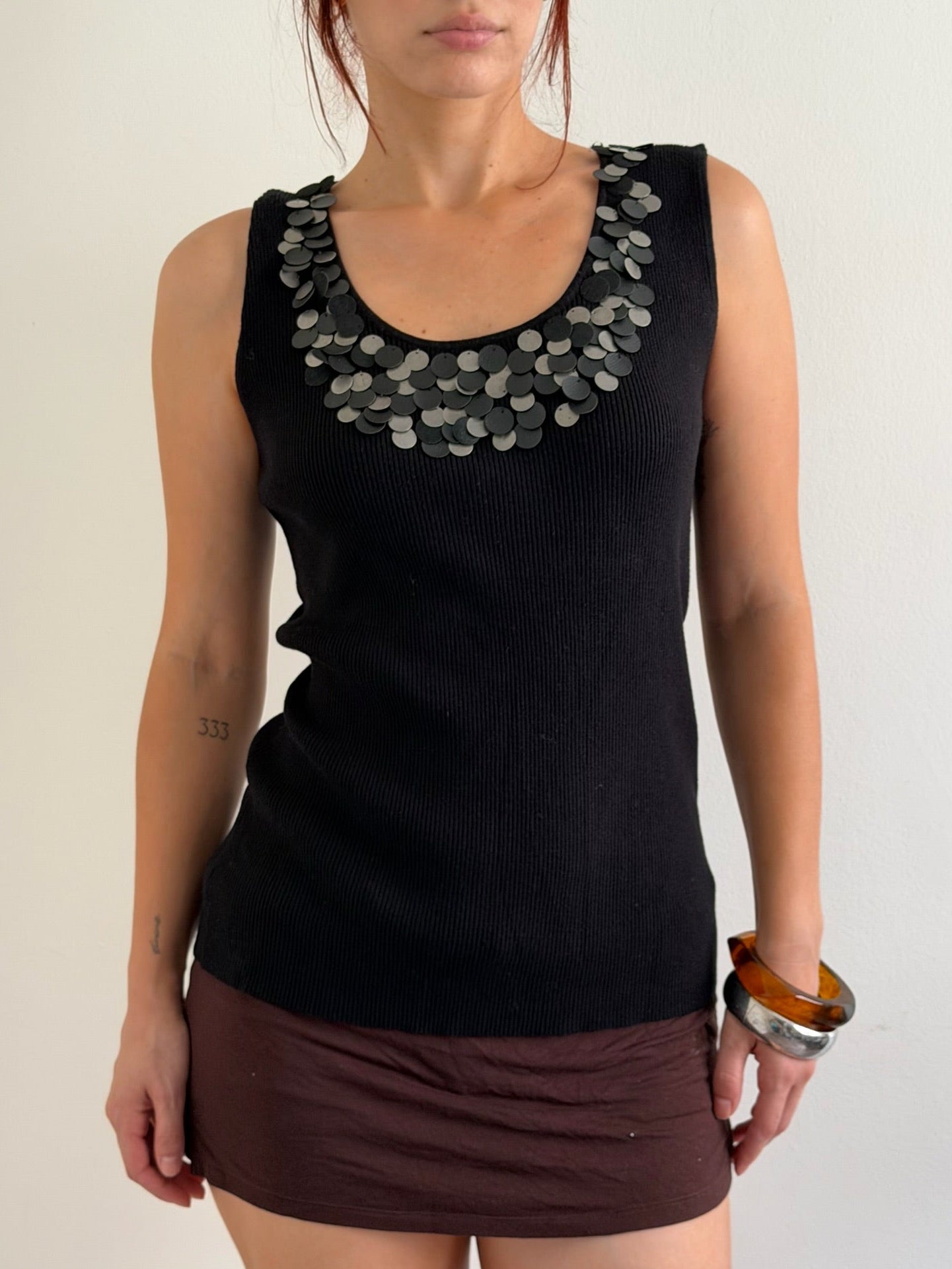 A woman wearing a black ribbed sleeveless top with a wide collar decorated with overlapping silver coin-like embellishments, paired with a dark brown mini skirt, against a plain light background.