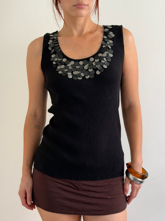 A woman wearing a black ribbed sleeveless top with a wide collar decorated with overlapping silver coin-like embellishments, paired with a dark brown mini skirt, against a plain light background.
