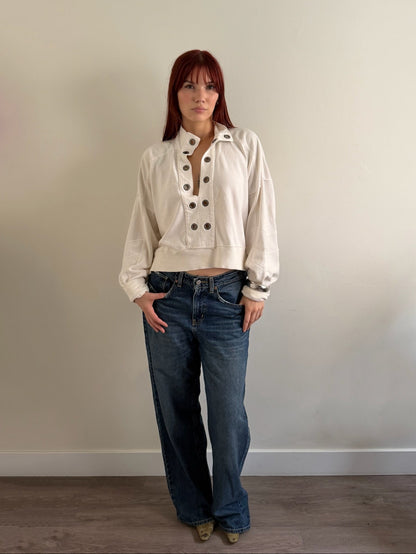 A woman with red hair stands against a plain beige wall wearing a cream-colored cropped button-up jacket with dark buttons, blue wide-leg jeans, and brown shoes.