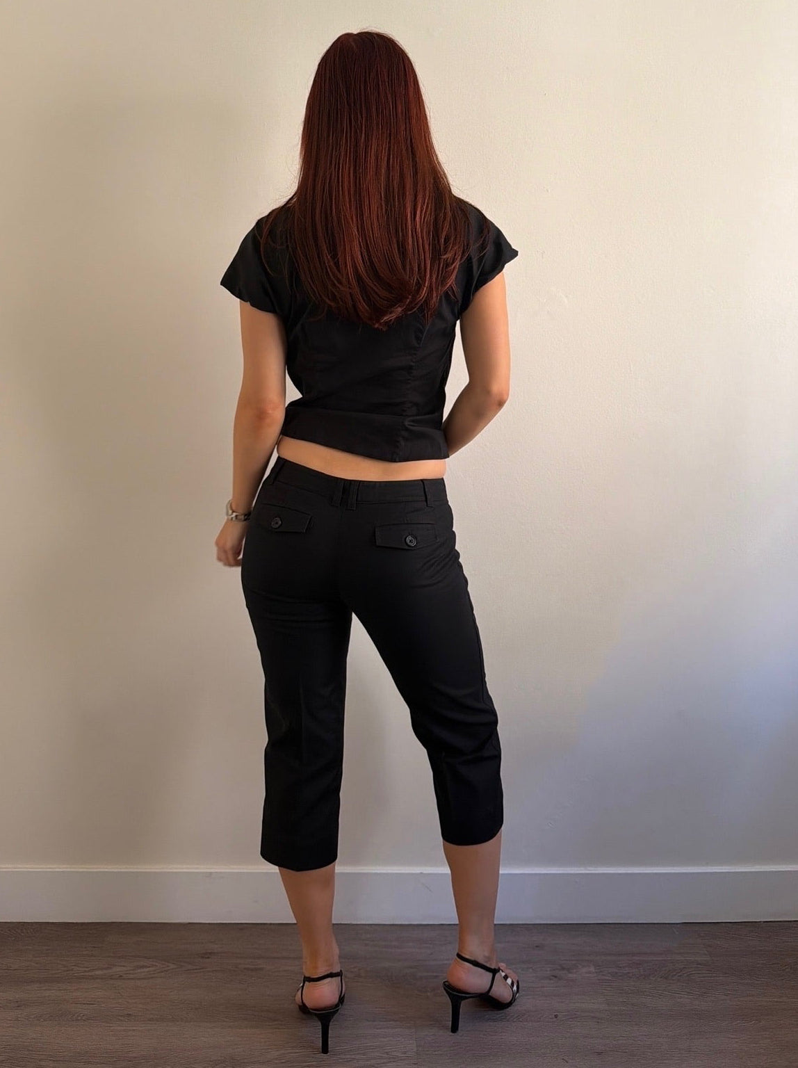 Person wearing a black crop top and black pants standing against a plain wall.