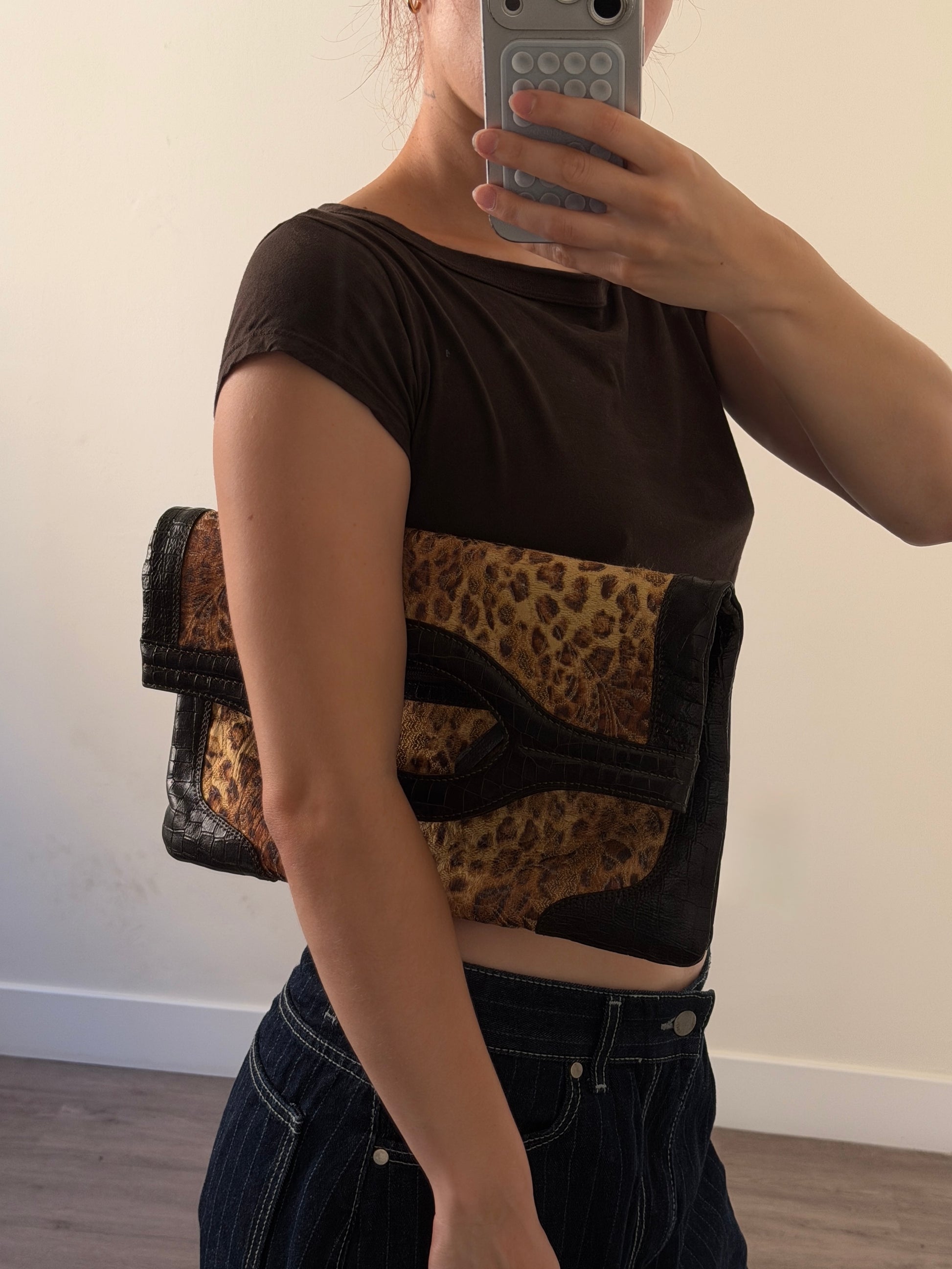 A person taking a mirror selfie wearing a dark brown short-sleeve crop top with a leopard-print and black fabric panel across the chest, paired with dark denim jeans. A leopard-print and black shoulder bag is slung over the shoulder.