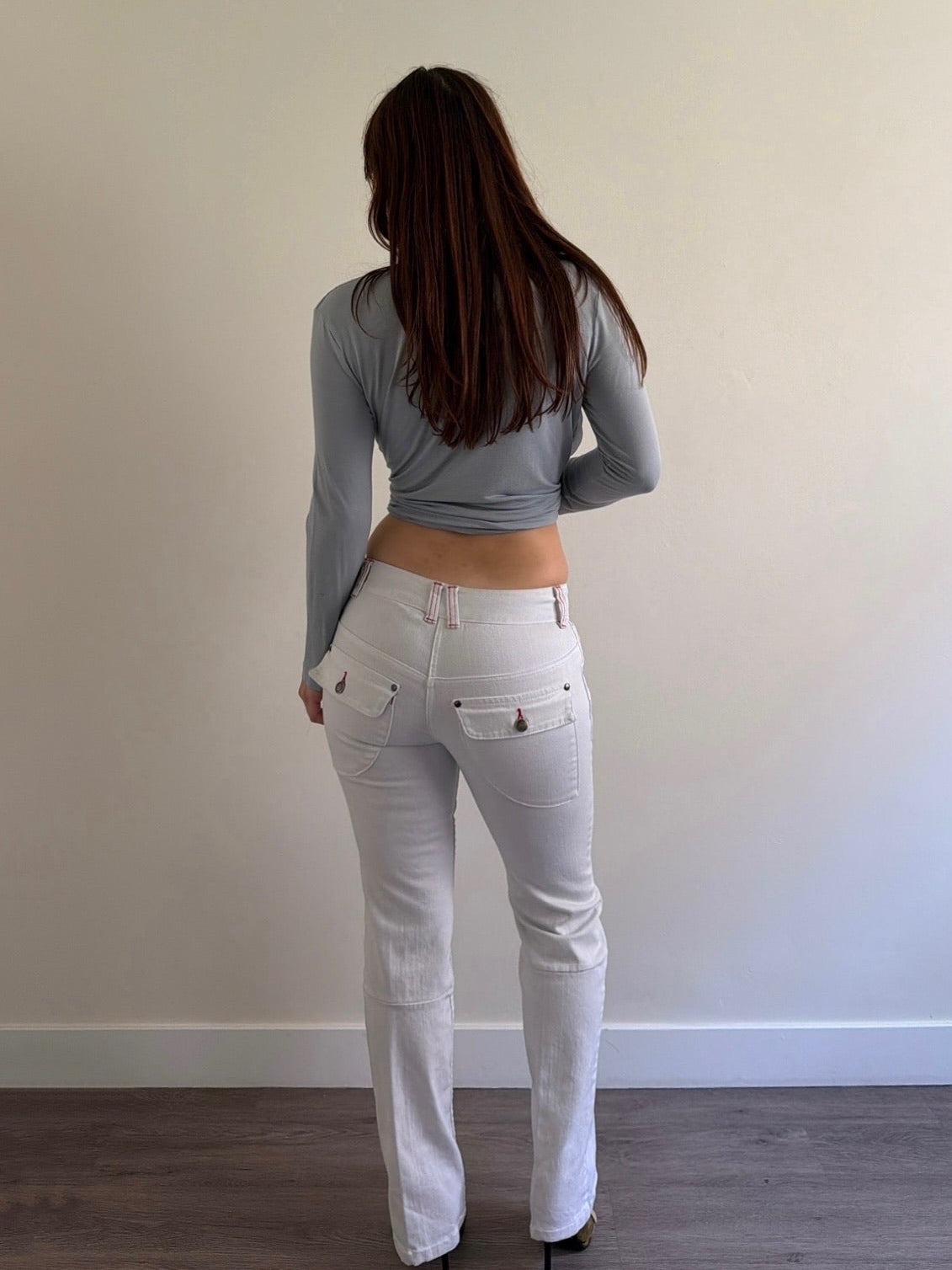 Person wearing a gray long-sleeve top and white pants standing against a plain wall.