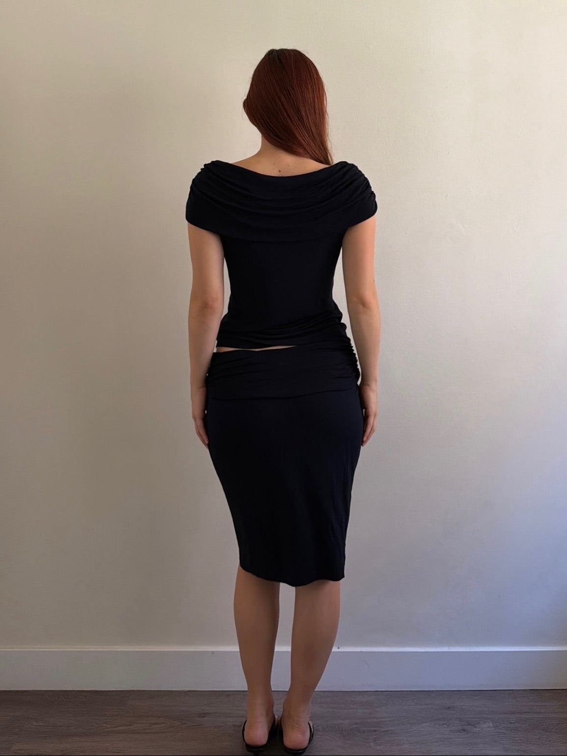 A woman with red hair is shown from behind wearing a black draped short-sleeve top and matching black knee-length skirt, standing against a plain beige wall.