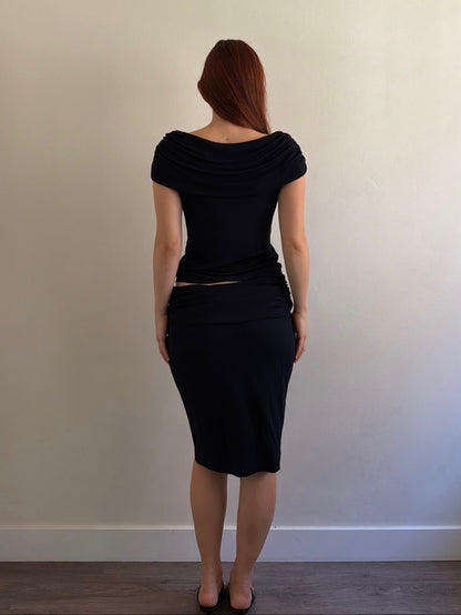 A woman with red hair is shown from behind wearing a black draped short-sleeve top and matching black knee-length skirt, standing against a plain beige wall.