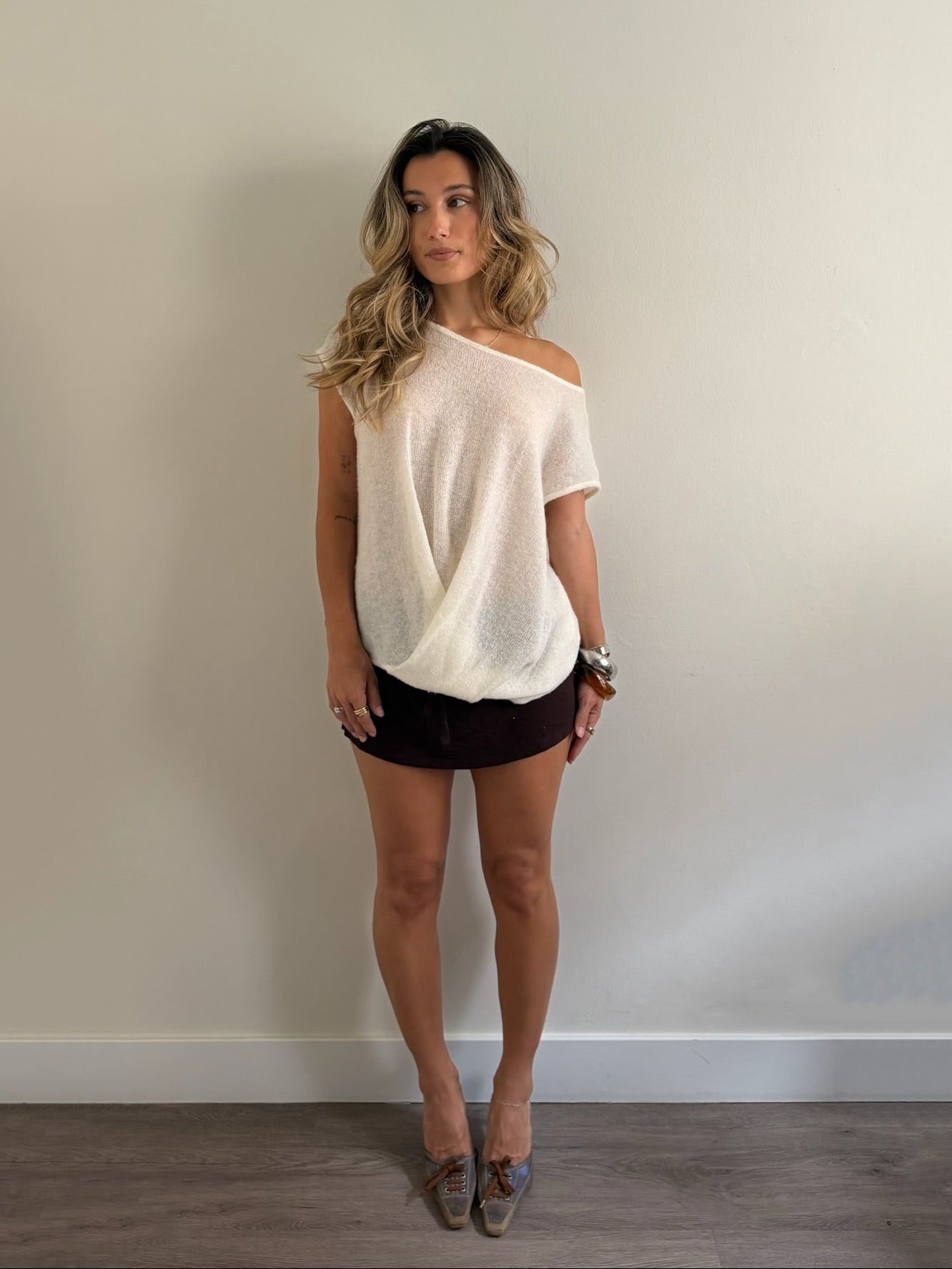 A woman stands against a plain beige wall wearing a cream off-the-shoulder knit top, dark brown shorts, and gray loafers.