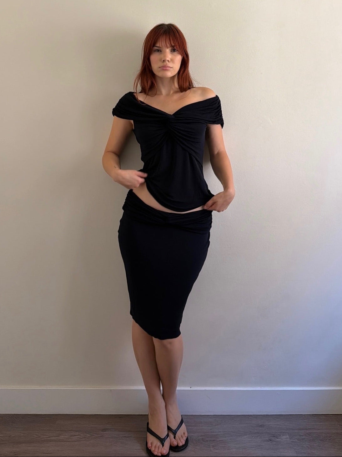 A woman with red hair wearing a black off-the-shoulder top and matching black pencil skirt, standing against a plain beige wall.