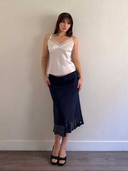 Woman wearing a white top and navy skirt against a plain wall.