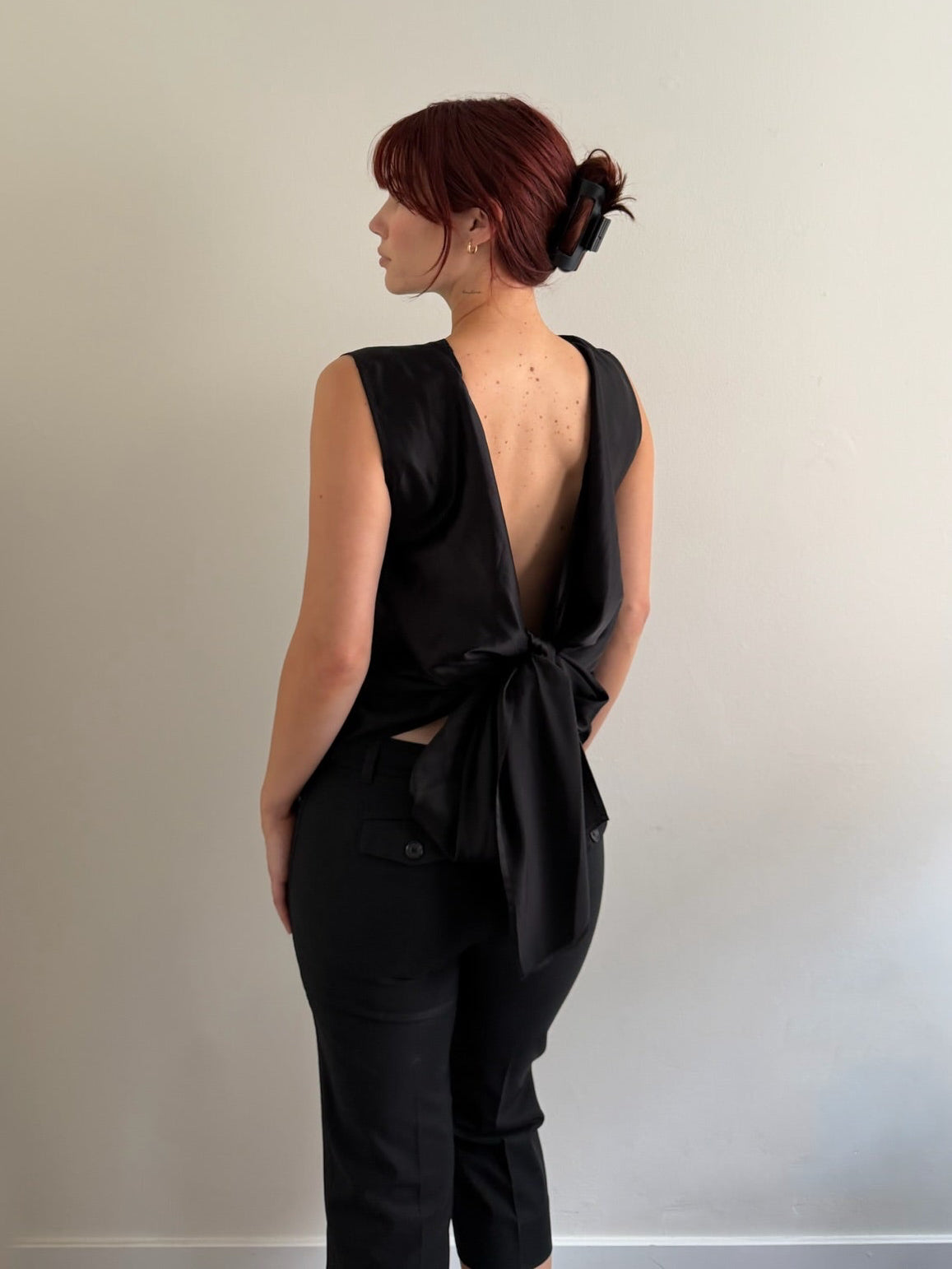 A woman with red hair tied up in a bun is shown from the back, wearing a black sleeveless top with a deep open back and a large bow detail, paired with black pants, standing against a plain light gray wall.