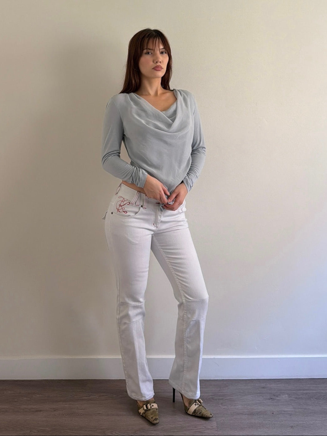 Woman wearing a gray sweater and white jeans against a plain wall.