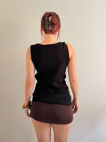 A woman viewed from behind wearing a sleeveless black top and a brown mini skirt, standing against a plain light gray wall.