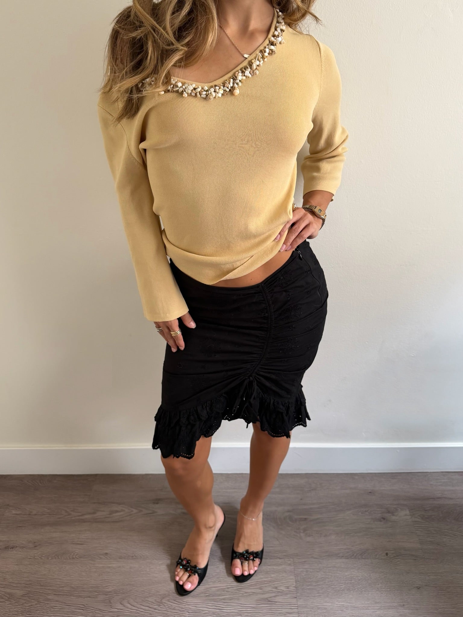 A woman modeling a beige knit long-sleeve top with a beaded neckline and a black ruched mini skirt, paired with black slide sandals, standing against a plain off-white wall.