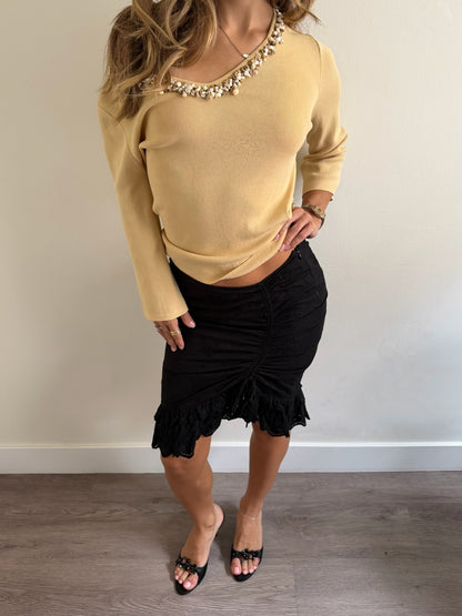 A woman modeling a beige knit long-sleeve top with a beaded neckline and a black ruched mini skirt, paired with black slide sandals, standing against a plain off-white wall.