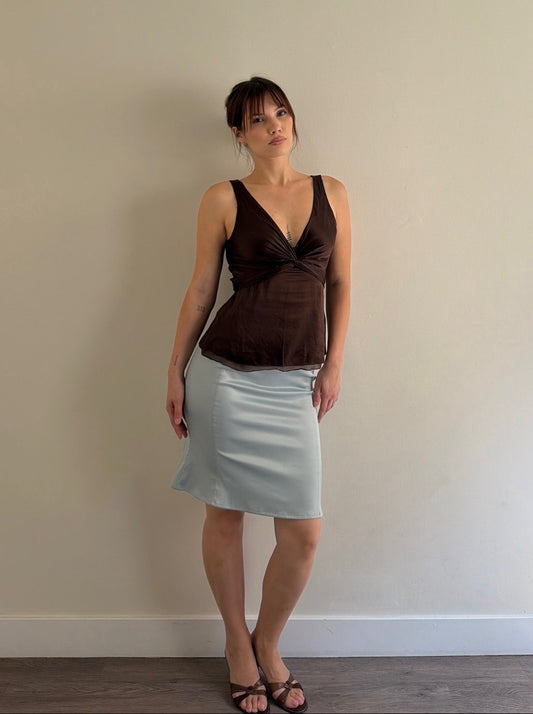 Woman wearing a brown top and light blue skirt against a plain wall.
