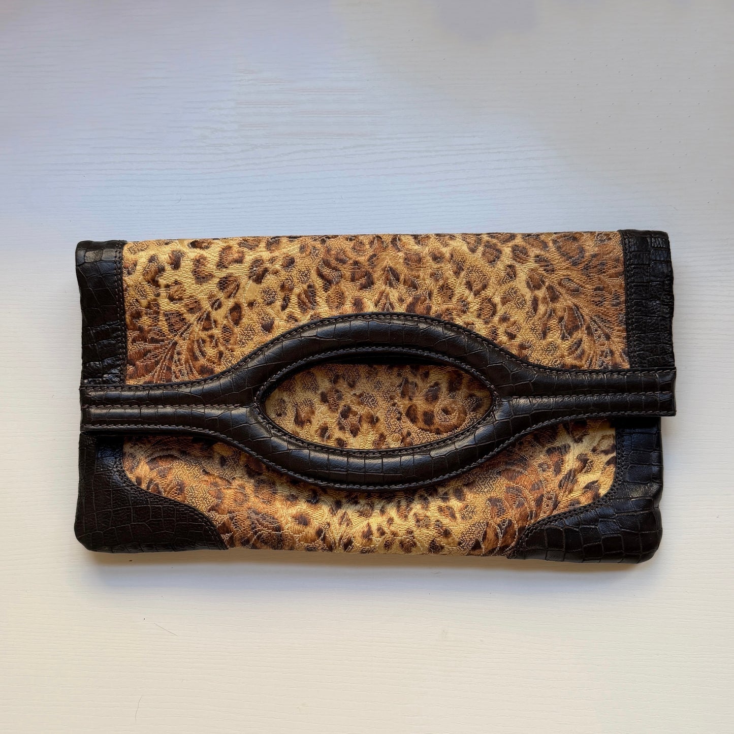 A leopard print clutch wallet with dark brown faux-croc trim and a central oval handle cutout, shown on a light off-white background.