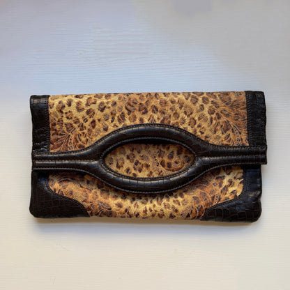 A leopard print clutch wallet with dark brown faux-croc trim and a central oval handle cutout, shown on a light off-white background.