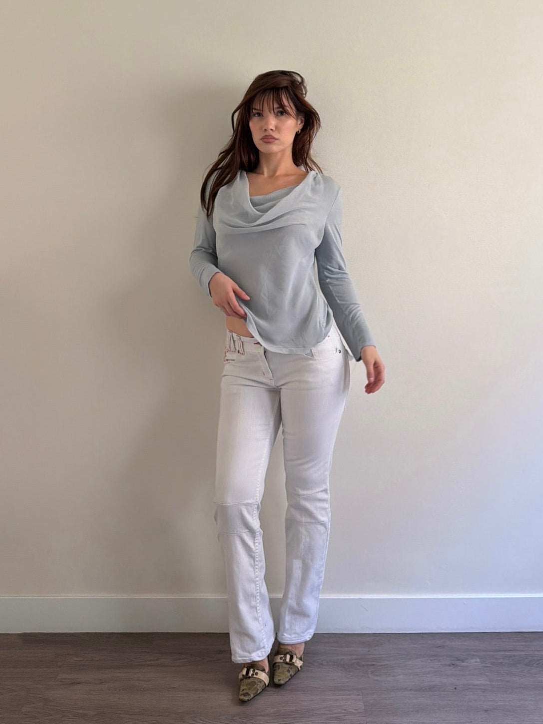 Woman wearing a light gray long-sleeve top and white jeans against a plain wall.